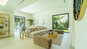 A modern living room with a sofa, ottoman, and coffee table, large windows, and a decorative wall piece. - Magnificient 5 bed villa in Golden Mile Marbella (Marbella)