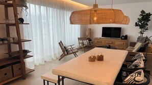 Un salon moderne avec un coin repas, comprenant une table en bois, des chaises, un téléviseur et une grande fenêtre agrémentée de rideaux légers.