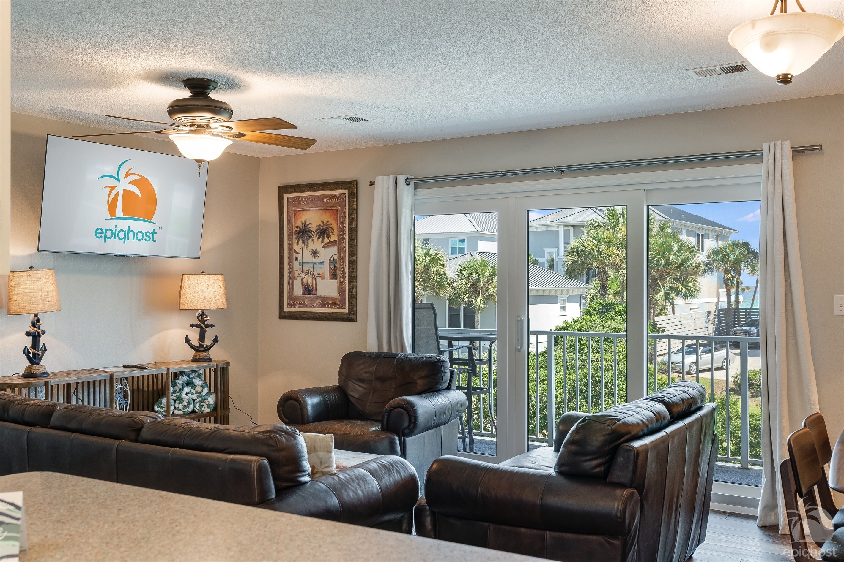 A spacious living area with a ceiling fan, a large TV displaying the 'epighost' logo, a framed artwork, and a balcony with a view of palm trees and houses.