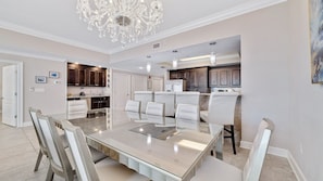 A spacious dining area with a large glass table and white chairs, a kitchen with dark wooden cabinets, and a chandelier.
