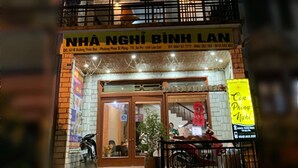 Un edificio de varios pisos con una fachada luminosa y faroles de colores. La planta baja muestra un letrero que dice "NHÀ NGHÍ BINH LAN" con datos de contacto.
