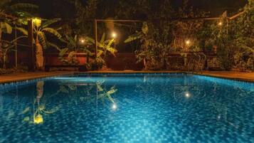 Piscina à noite com piso de azulejos e vegetação ao redor.