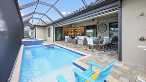 Ein Pool mit Fliesen umgeben, mit blauen Liegestühlen, eine Terrasse mit Esstisch und Stühlen sowie ein Blick in den Garten durch eine Glastür.