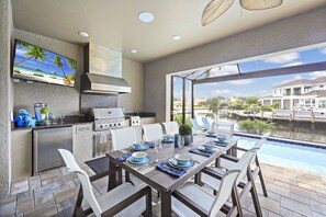 A modern outdoor kitchen with a dining area overlooking a water body. - Waterfront Home | W Exposure, Pool & Spa (Marco Island)
