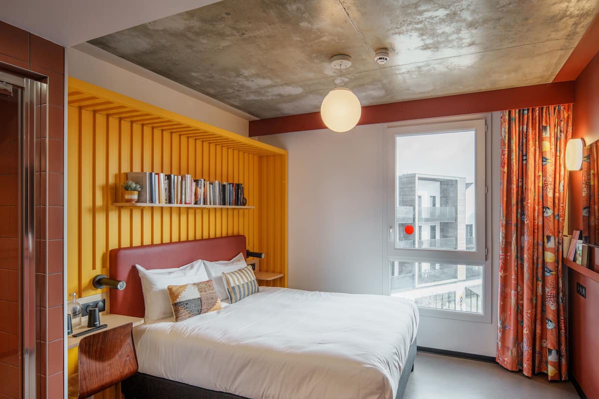 A modern hotel room with a large bed, a bookshelf, a window with a city view, and a unique ceiling design.