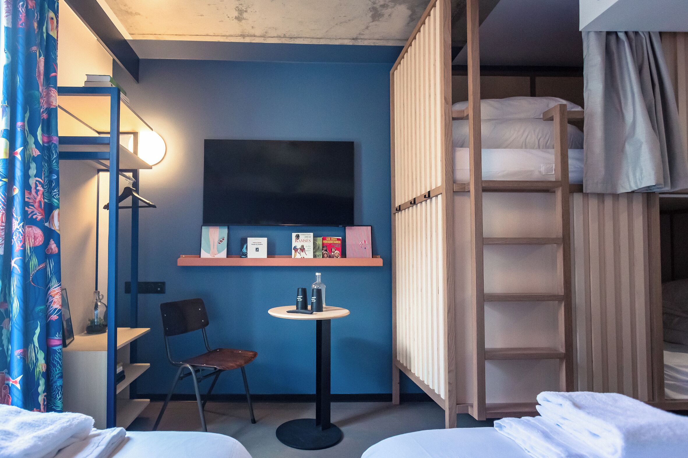 A room with bunk beds, a small table with a lamp, a chair, and a flat-screen TV mounted on the wall.