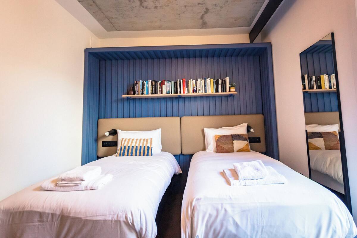 A hotel room with two beds, a bookshelf, and a mirror.