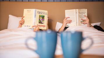 Two people reading books on a bed with two blue mugs in front of them.