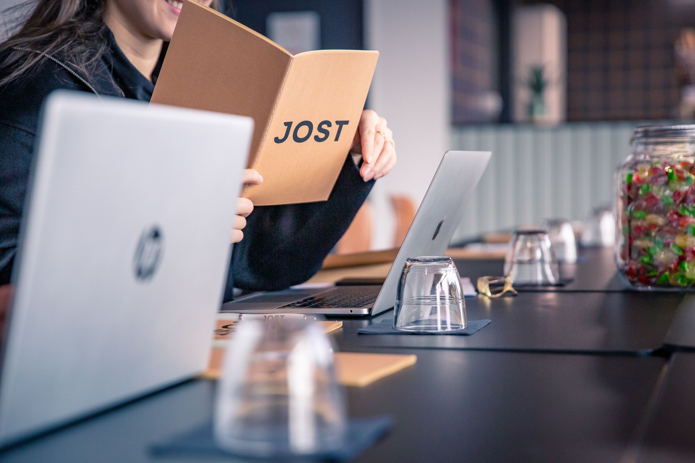 Una persona sostiene un libro titulado 'JOST' sobre una mesa con dos computadoras portátiles, un vaso de agua y un frasco de caramelos.