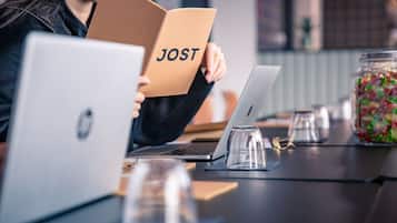 Una persona sostiene un libro titulado 'JOST' sobre una mesa con dos computadoras portátiles, un vaso de agua y un frasco de caramelos.