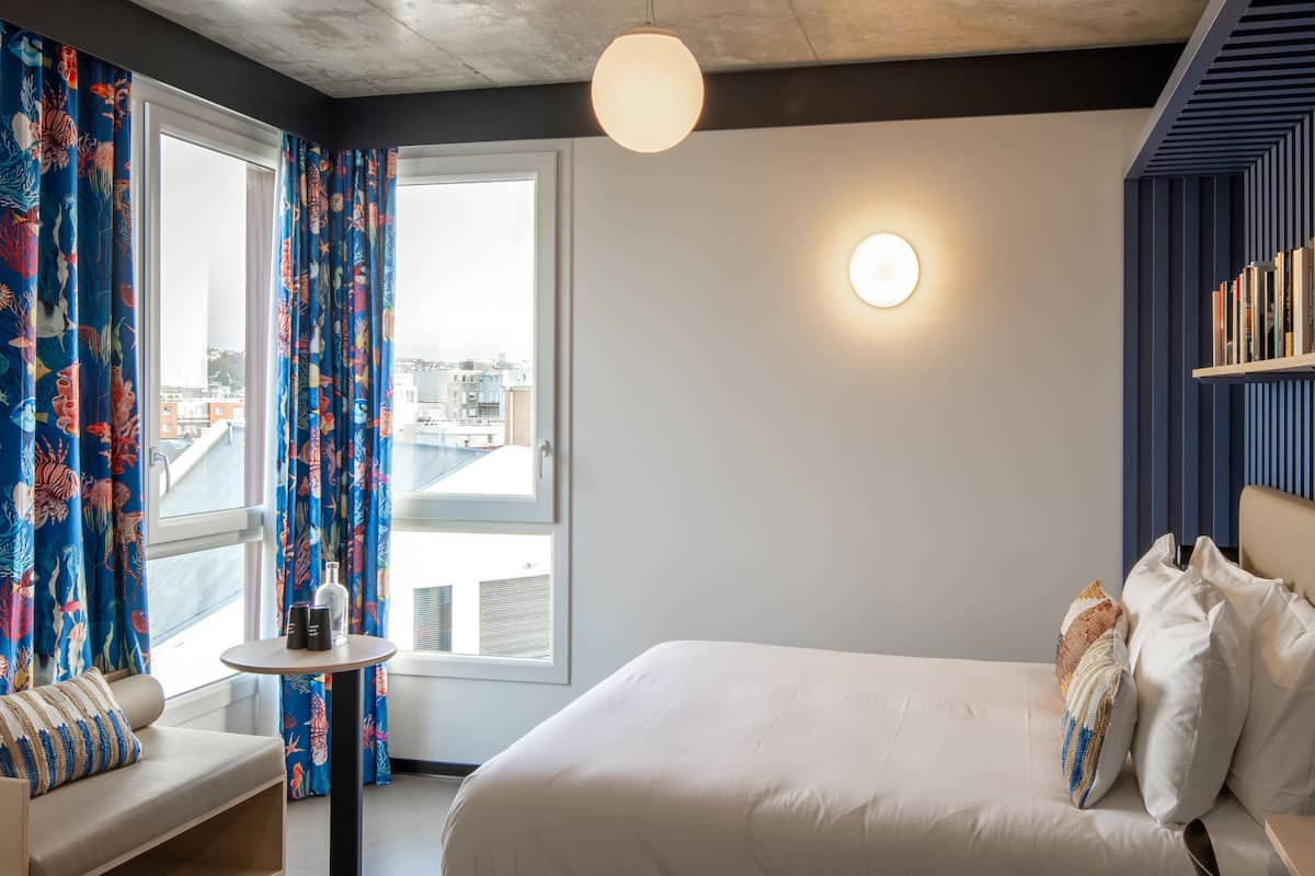 A hotel room with a bed, a small table, a sofa, curtains with a marine theme, a round light fixture, and a bookshelf.