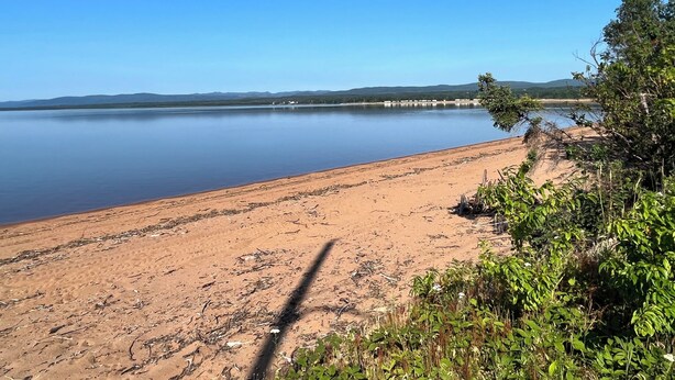 A sandy beach with a calm lake, surrounded by greenery and distant hills.