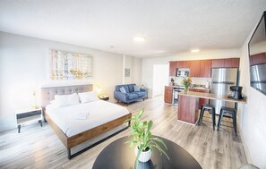 A modern hotel room with a large bed, a small sofa, a dining area with a kitchen, and a potted plant. - Mega Group Block: 4 Units | Sleeps 10-16 (Deerfield Beach)
