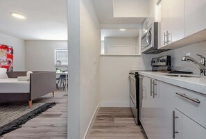 Una cocina moderna con armarios blancos, un microondas y un lavamanos. En la habitación contigua, hay una cama con cabecera gris y una alfombra negra.