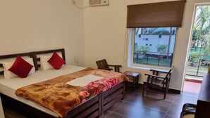 A bedroom with a wooden bed, a small desk, and a window with a view of a building and greenery. - Yoga Rishi (Narendranagar)