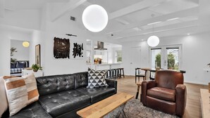 Una sala moderna con un sofá de cuero negro, una mesa de centro de madera y un sillón de cuero marrón. En el fondo, hay una zona de cocina con armarios blancos y una mesa de comedor.