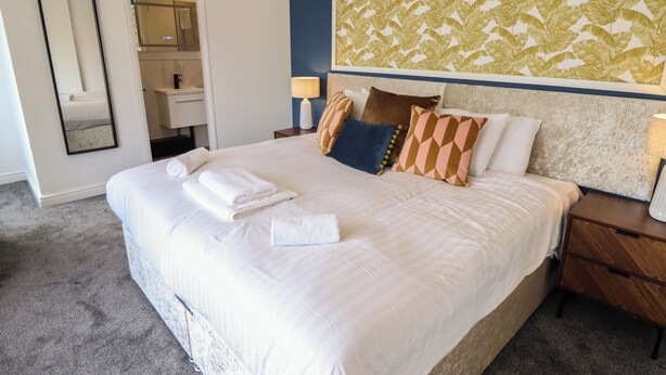 A neatly made bed with white linens, a headboard with a leaf pattern, and a nightstand with a lamp. - Carlton House (Skegness)