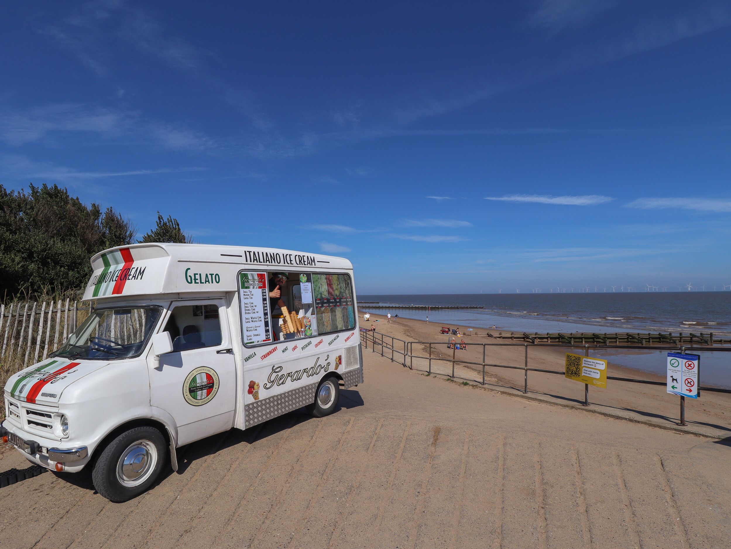 Un camión de helados estacionado junto a la playa, con vista al océano y turbinas eólicas al fondo.
