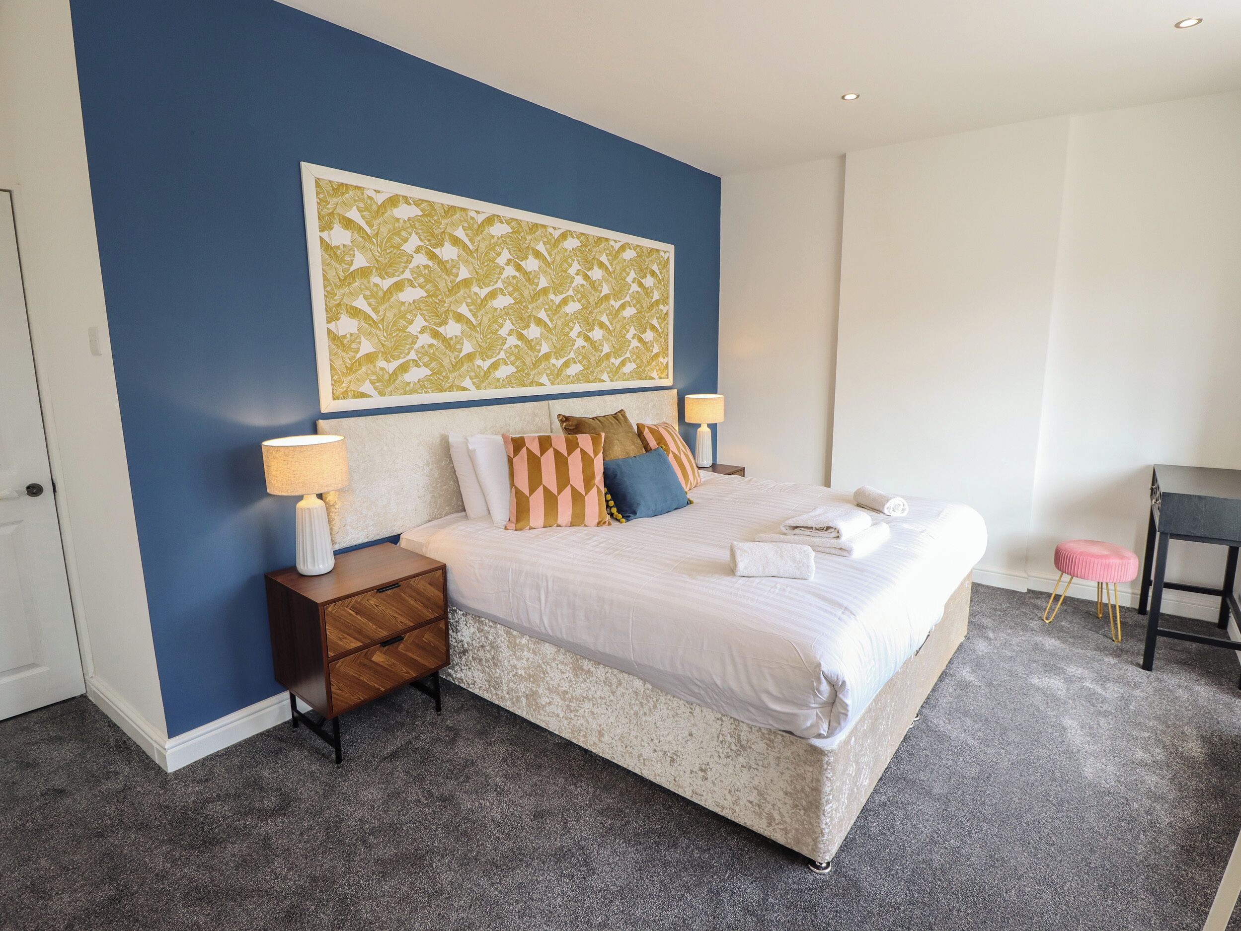 A neatly made bed with pillows and a headboard, a bedside table with a lamp, and a small stool.