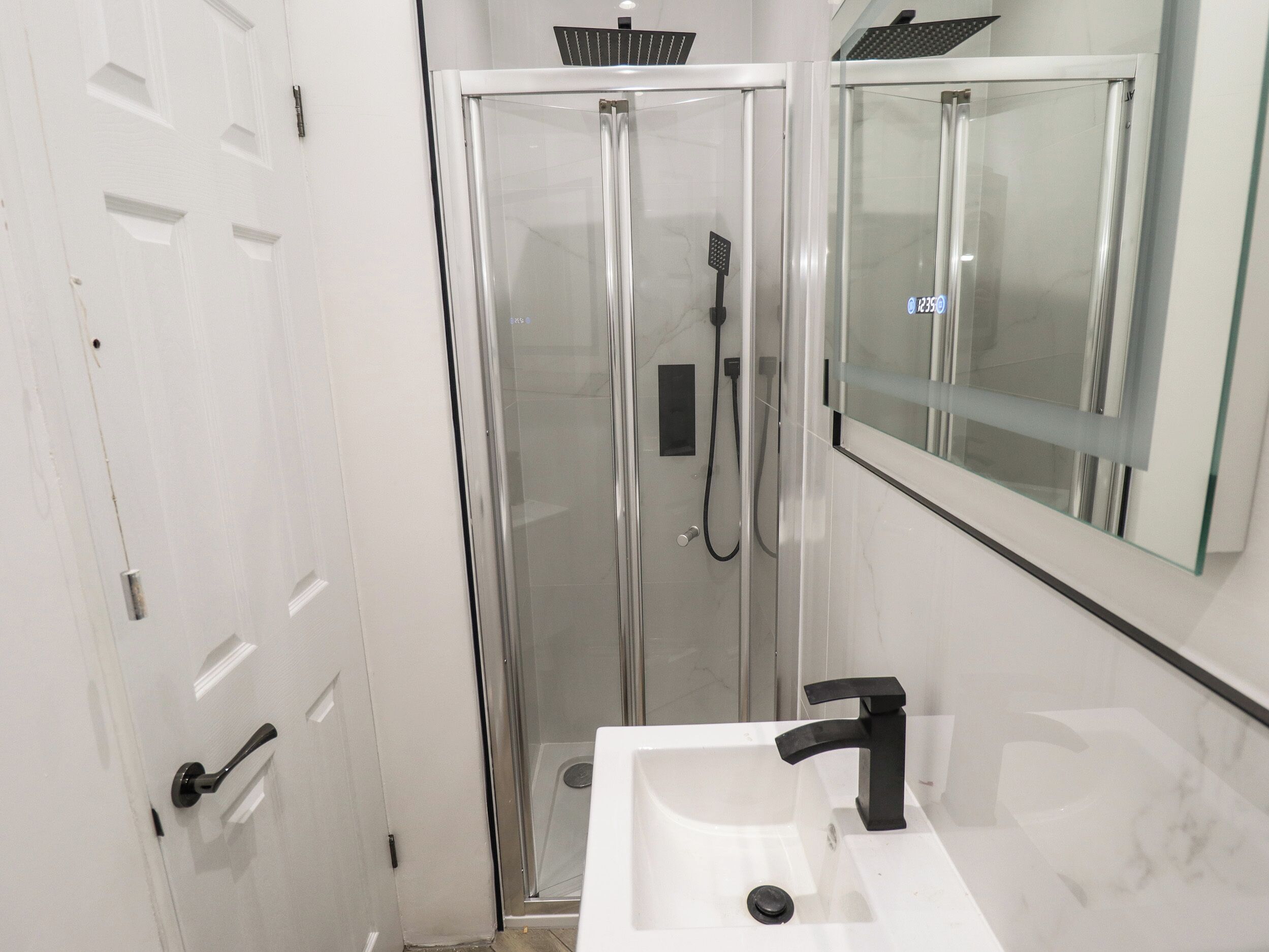 Un baño moderno con ducha acristalada, lavabo blanco con grifo negro y espejo sobre el lavabo.