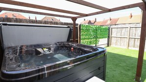 A hot tub with a wooden frame and a bench, set on artificial grass under a wooden pergola. - Carlton House (Skegness)