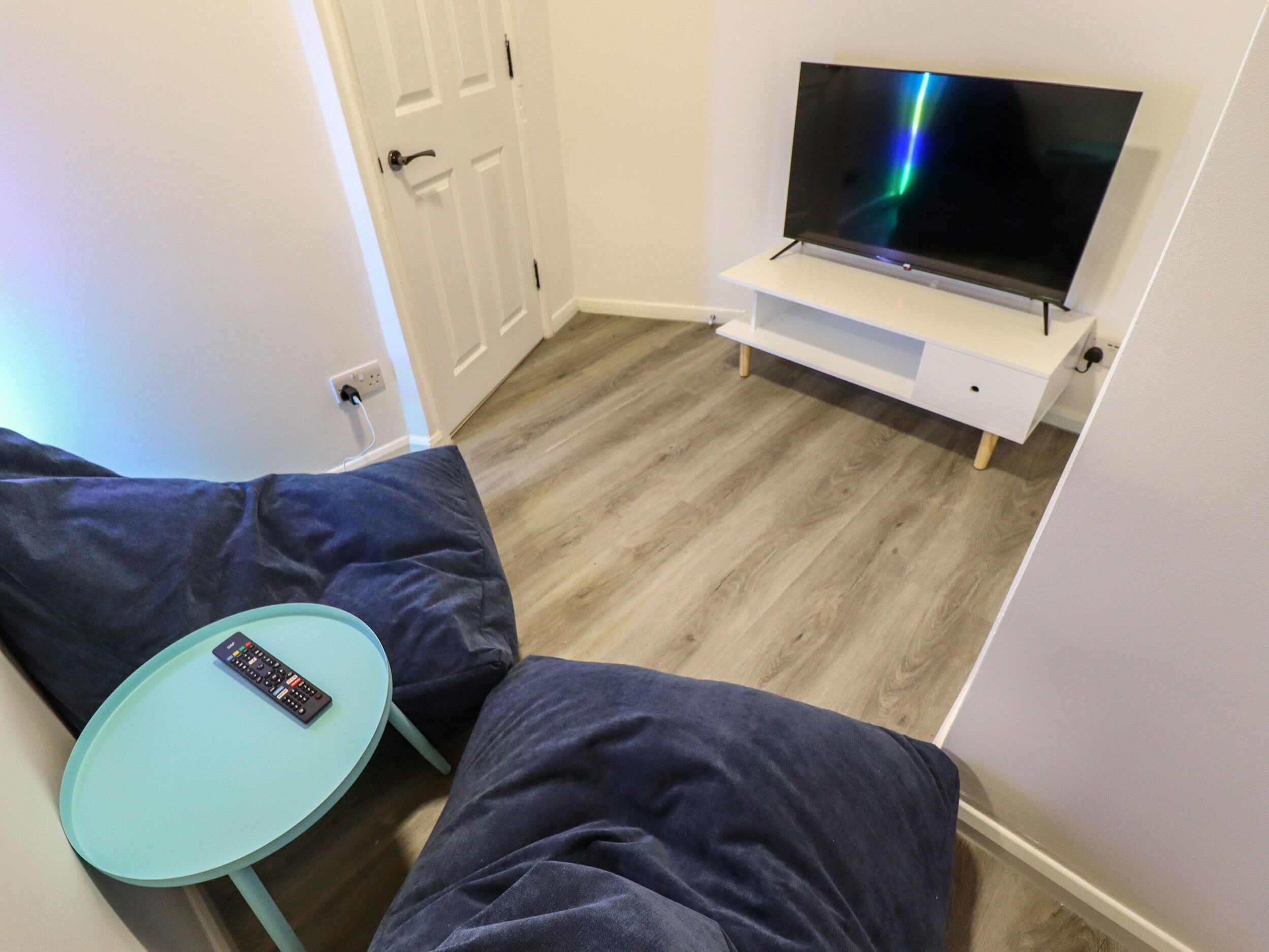 Um quarto pequeno com uma TV sobre um móvel, uma mesa redonda pequena e dois sofás escuros.