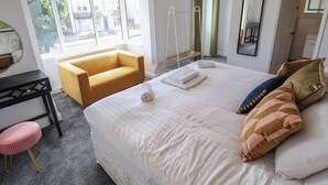A hotel room with a bed, a yellow armchair, a small stool, a mirror, and a window with curtains. - Carlton House (Skegness)