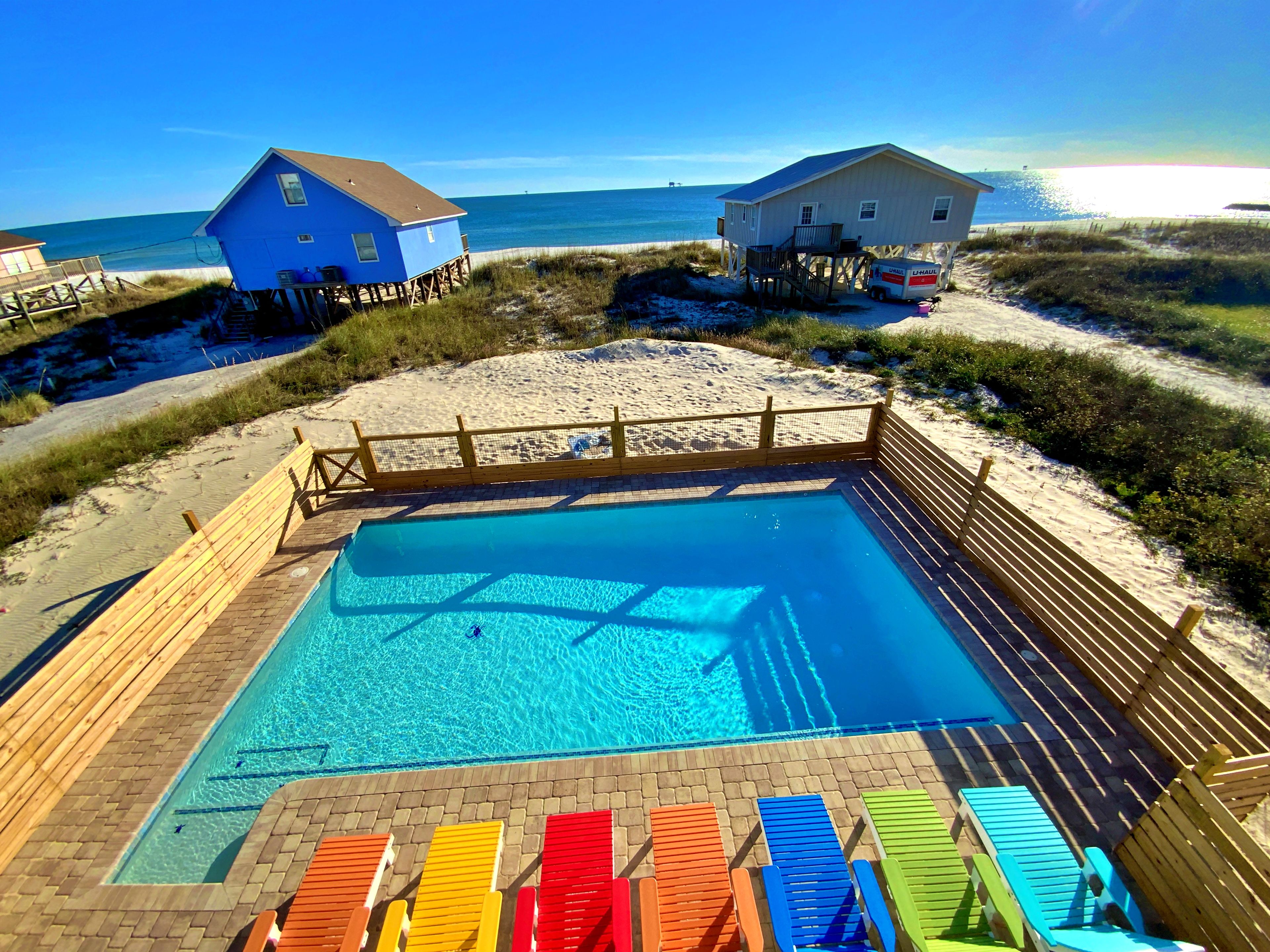 Una piscina con cubierta de madera y sillas de sol coloridas, rodeada por una valla de madera y dos casas de playa.