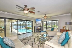 A spacious living room with a large glass door leading to a pool, ceiling fans, a television, and a bookshelf. - Southern Exposure Home W/ Expansive Canal Views! (Marco Island)