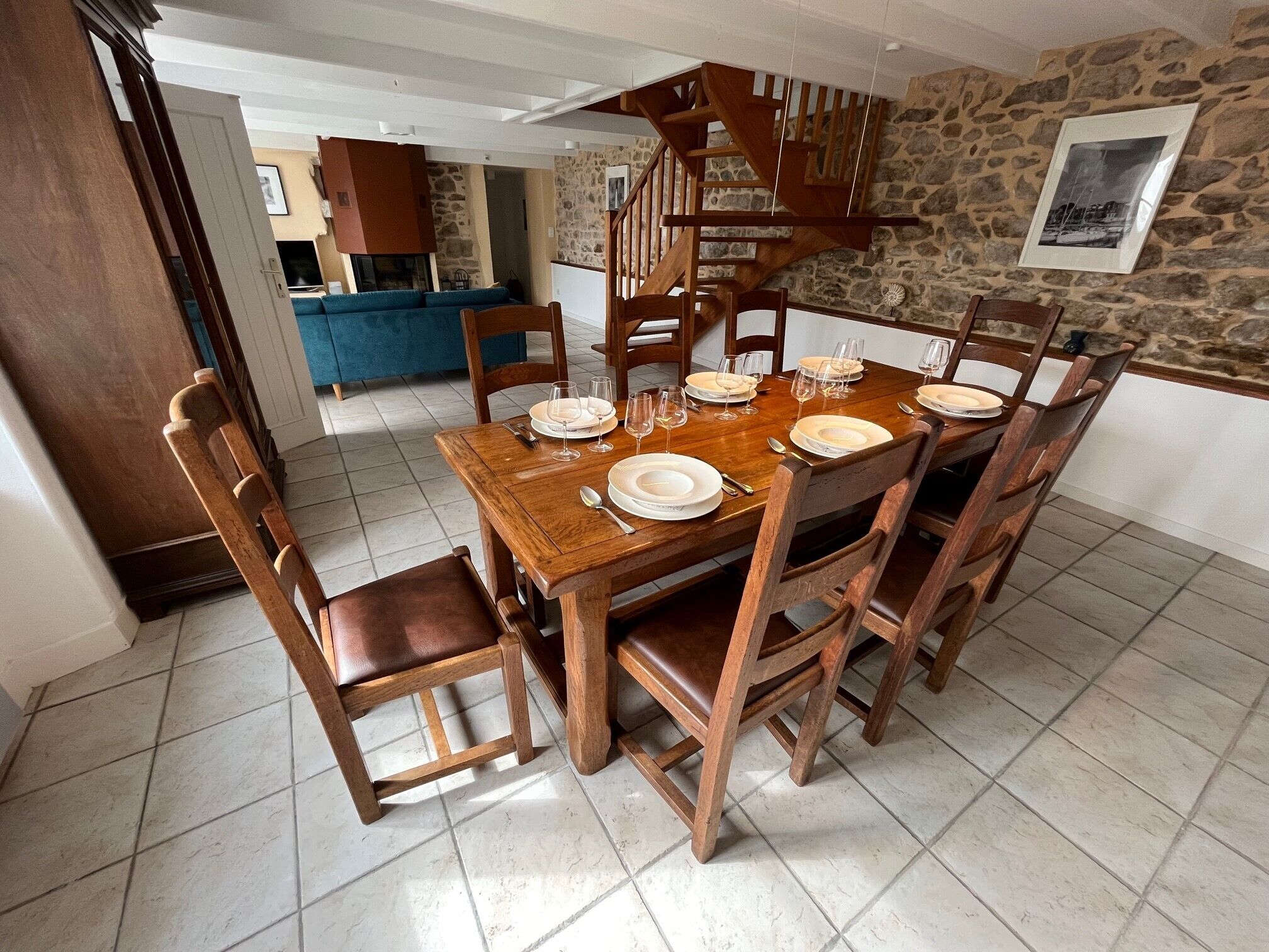 A dining area with a wooden table set for six, surrounded by wooden chairs. A stone wall and a wooden staircase are visible in the background.