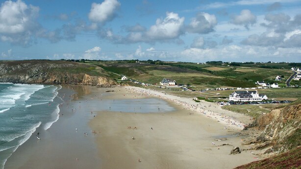 A coastal beach with a sandy shore, waves, and a grassy hillside with buildings.
