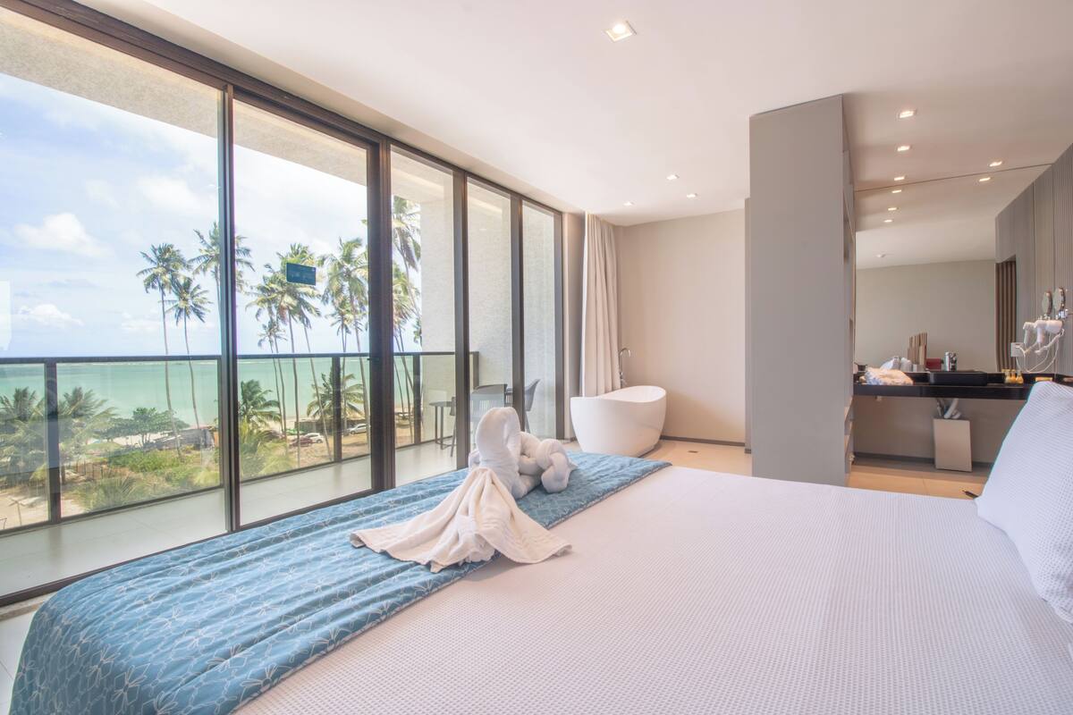 Una habitación de hotel moderna con una cama grande, un balcón con vistas a palmeras y al océano, un baño independiente y una pequeña kitchenette.