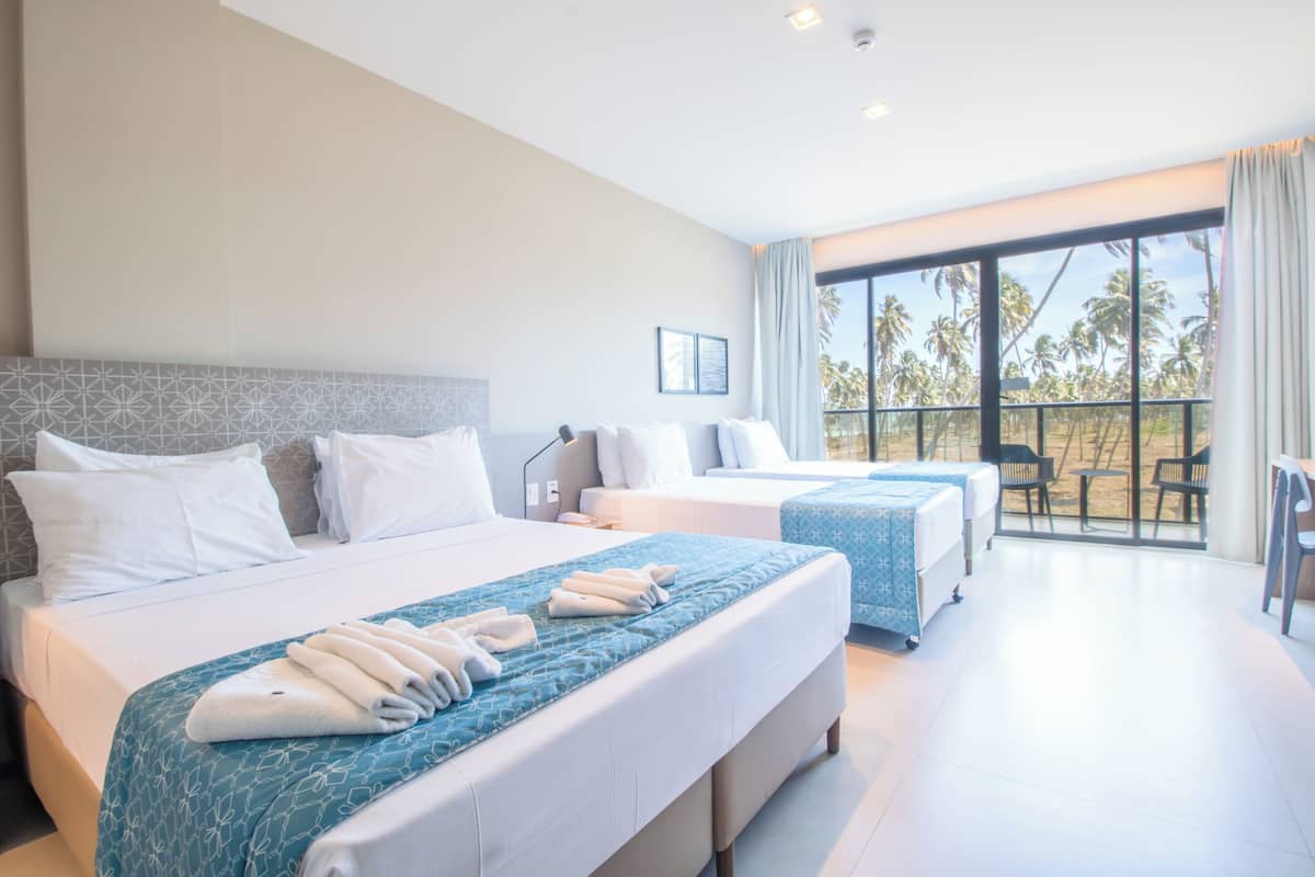 Habitación de hotel con dos camas, balcón con vista a palmeras y zona de estar con mesa y sillas.