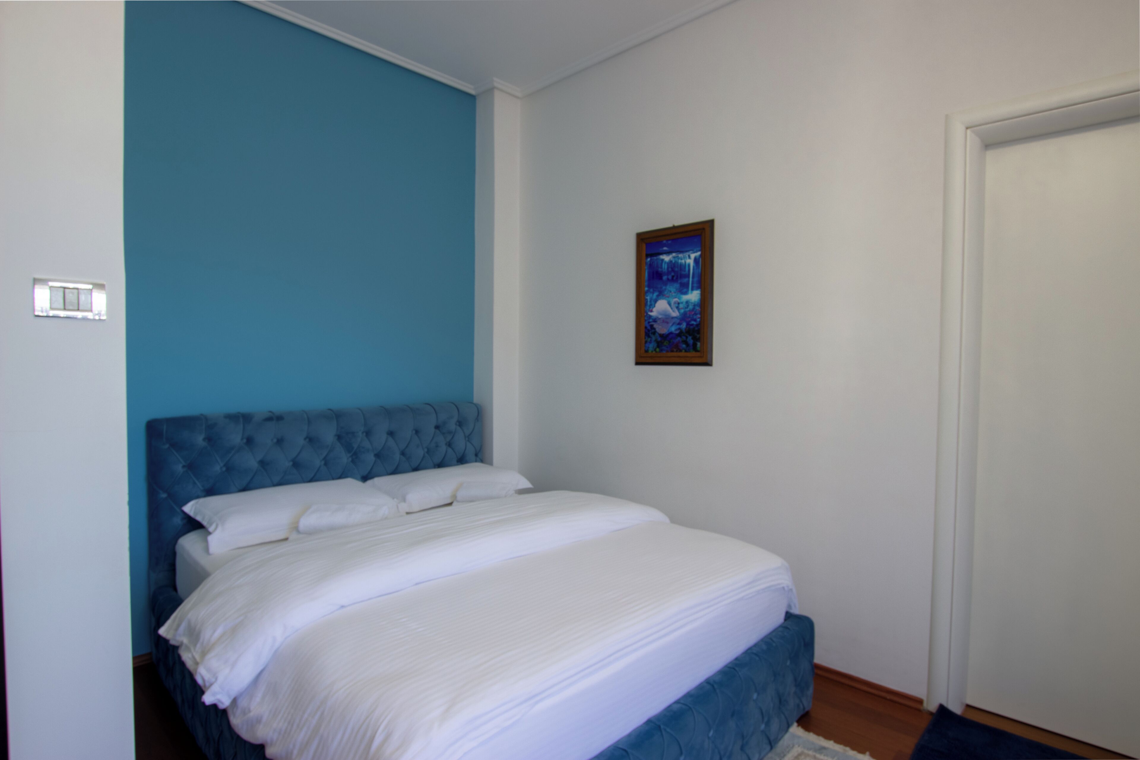 Una camera da letto con una testiera blu, biancheria da letto bianca, un quadro appeso al muro e una porta chiusa.