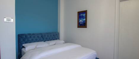 Una camera da letto con una testiera blu, biancheria da letto bianca, un quadro appeso al muro e una porta chiusa.