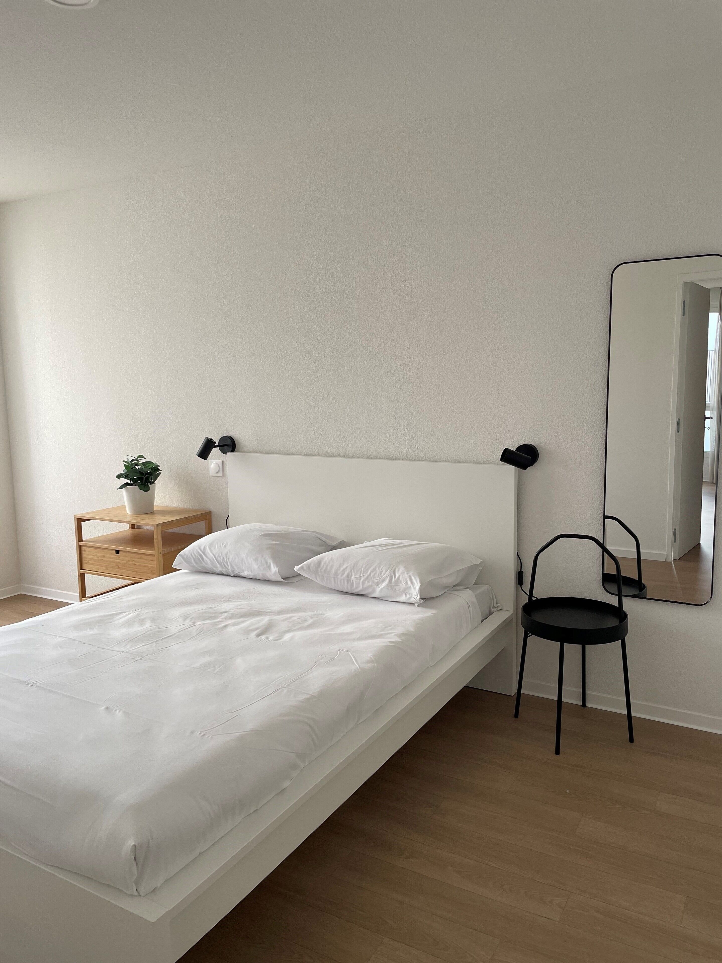Une chambre à coucher au style minimaliste, avec un lit simple, une table de chevet agrémentée d’une plante, une chaise et un miroir au mur.