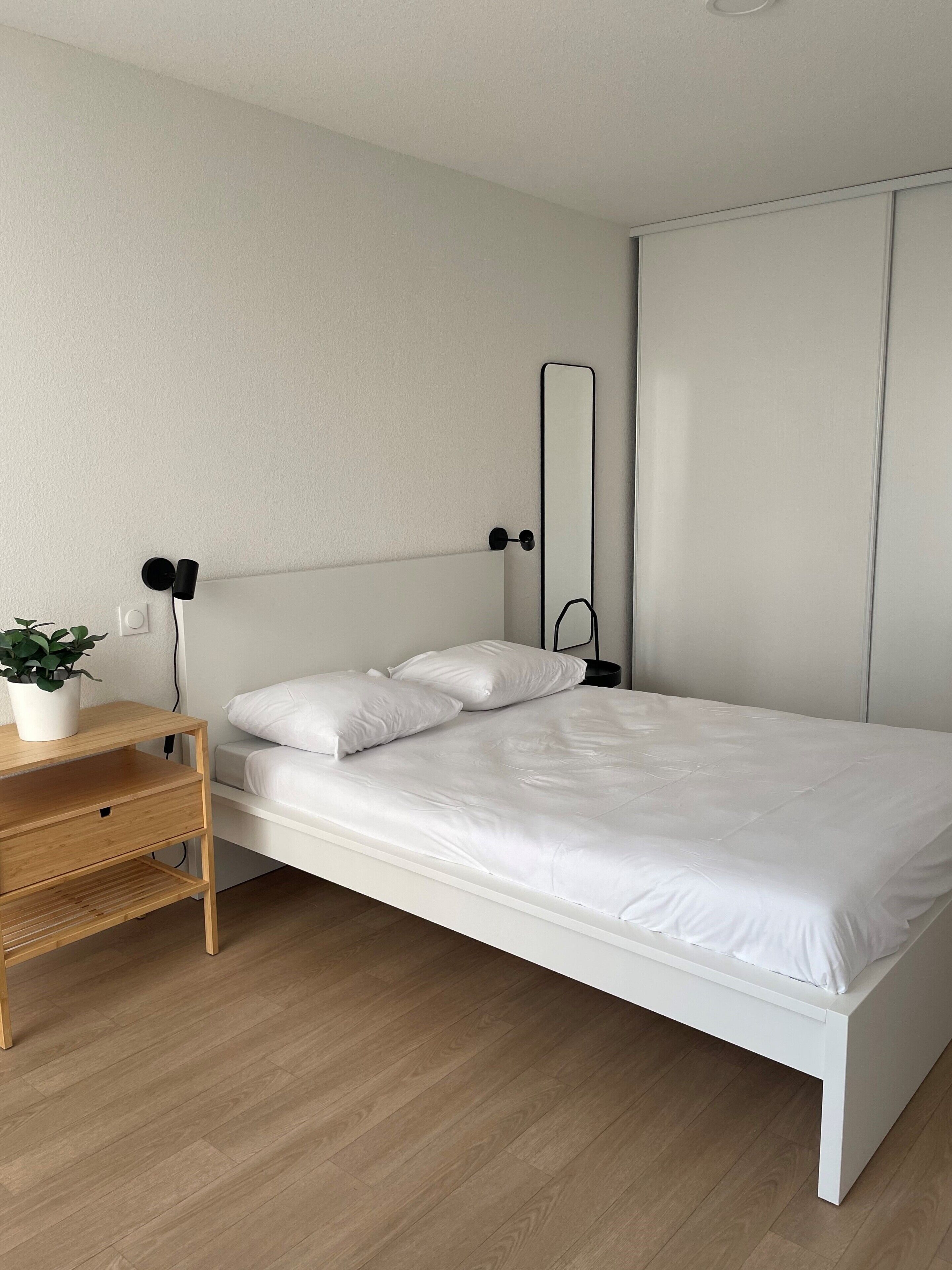 Quarto minimalista com cama branca, mesa de cabeceira de madeira, planta em vaso e um espelho grande na parede.