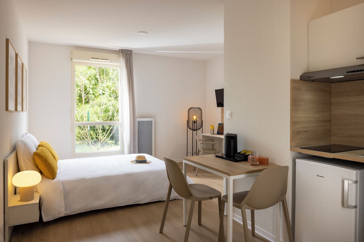 A modern hotel room with a bed, bedside table, lamp, and a view of greenery through the window.