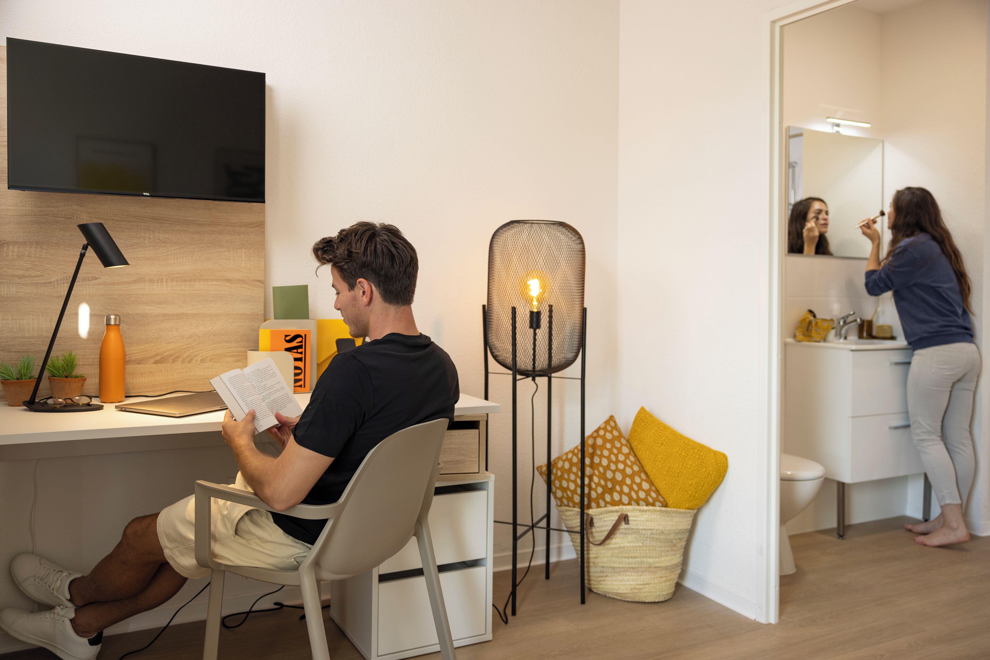 A modern room with a desk, chair, and a person reading a book. There's a TV mounted on the wall, a lamp, and a basket with yellow pillows.