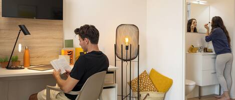 A modern room with a desk, chair, and a person reading a book. There's a TV mounted on the wall, a lamp, and a basket with yellow pillows.