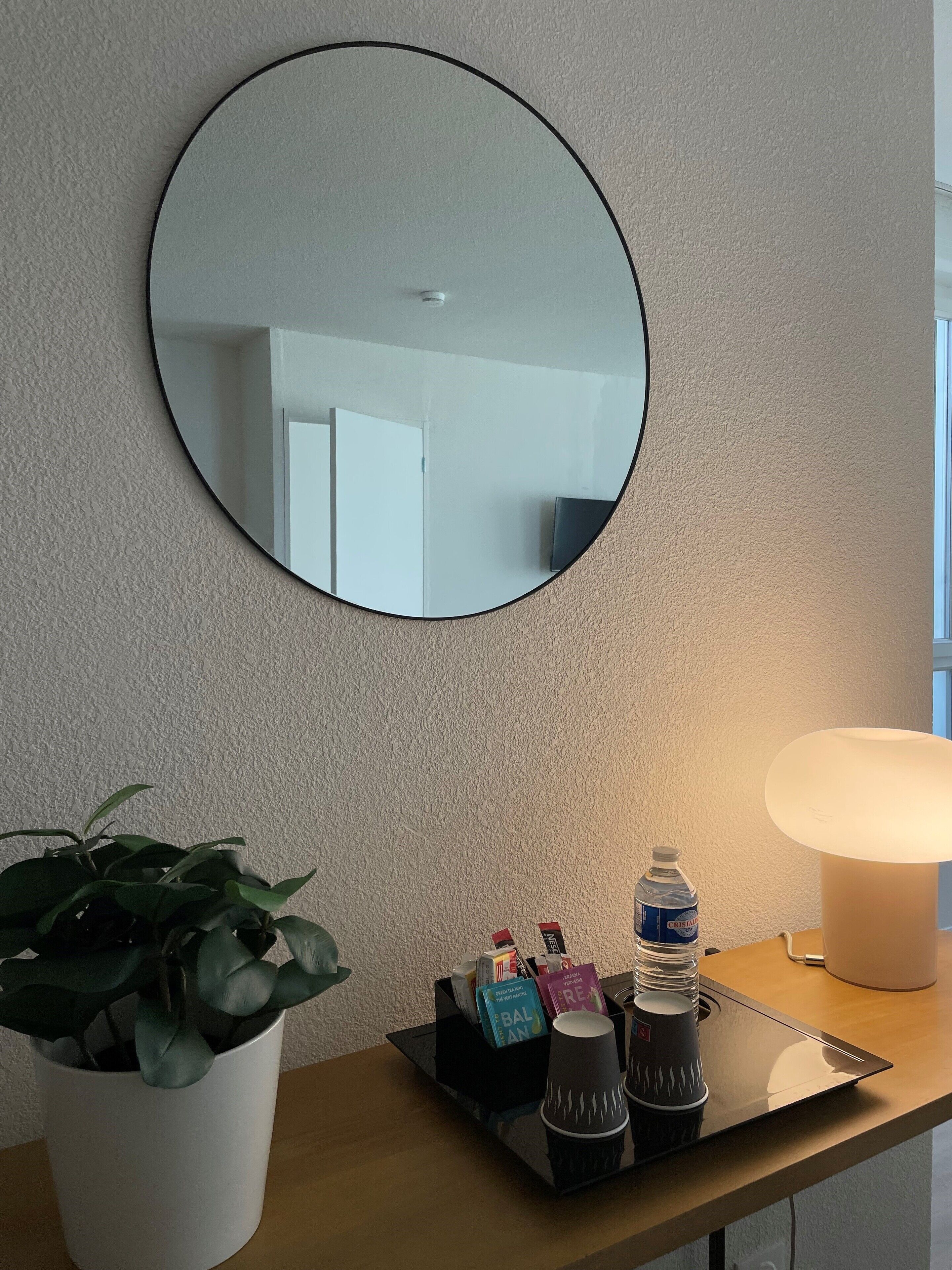 Habitación de hotel con un espejo redondo en la pared, una mesita de noche con lámpara, una planta en maceta y una bandeja con artículos de aseo y una botella de agua.