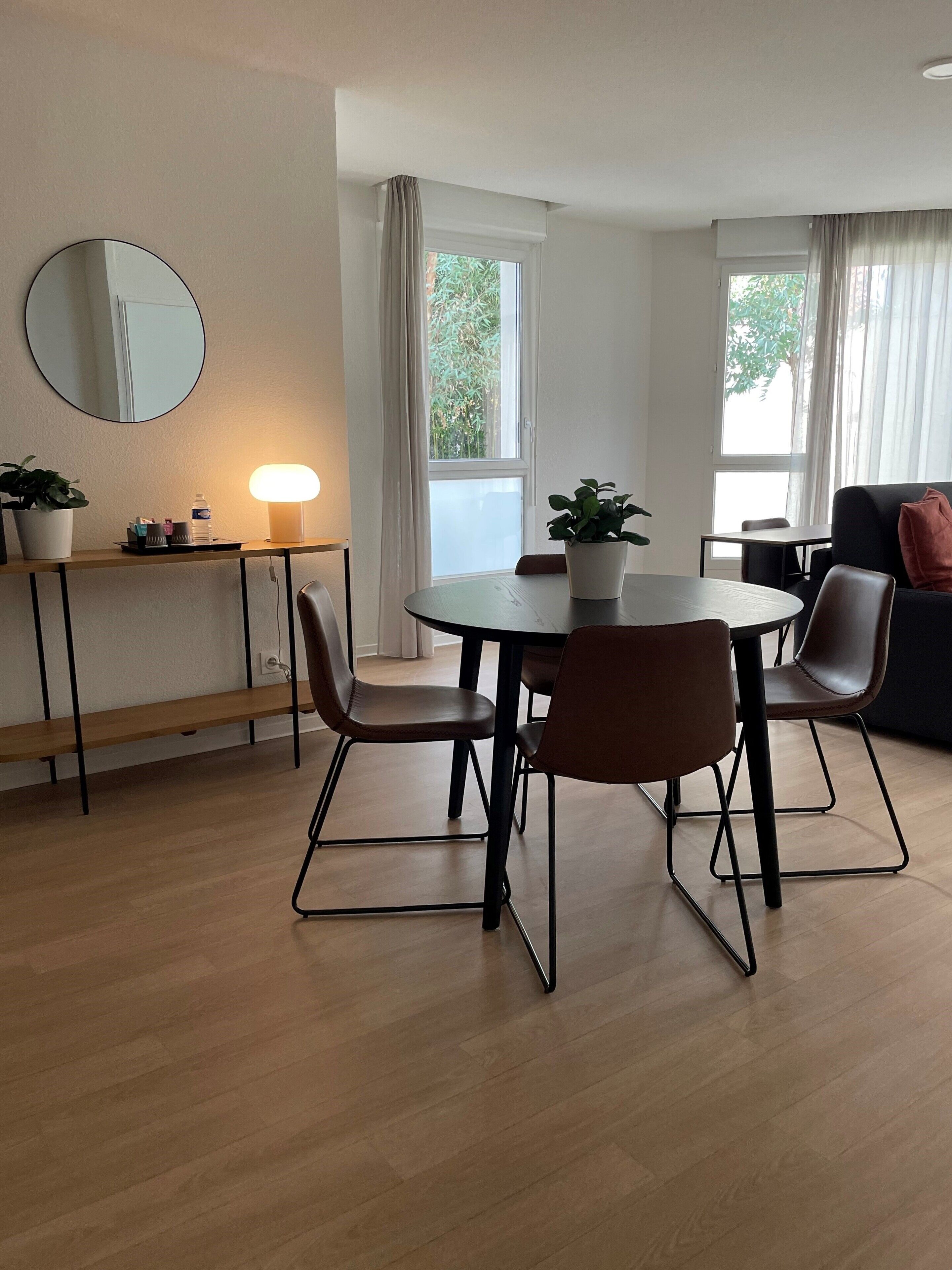 Una sala de estar moderna con una mesa redonda de comedor, dos sillas, un sofá, una mesa de centro, una mesita de noche con una lámpara, una planta en maceta y un gran ventanal con cortinas.