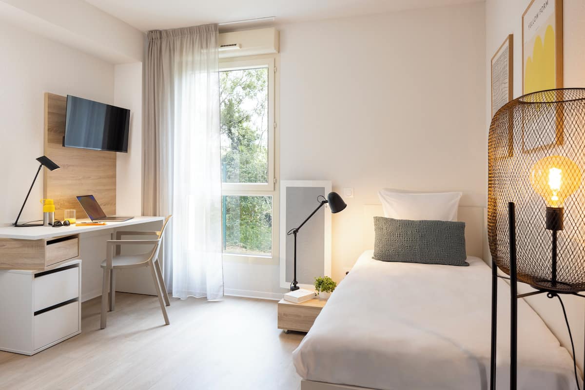 A modern hotel room with a desk, a bed, a TV mounted on the wall, and a large window with curtains.
