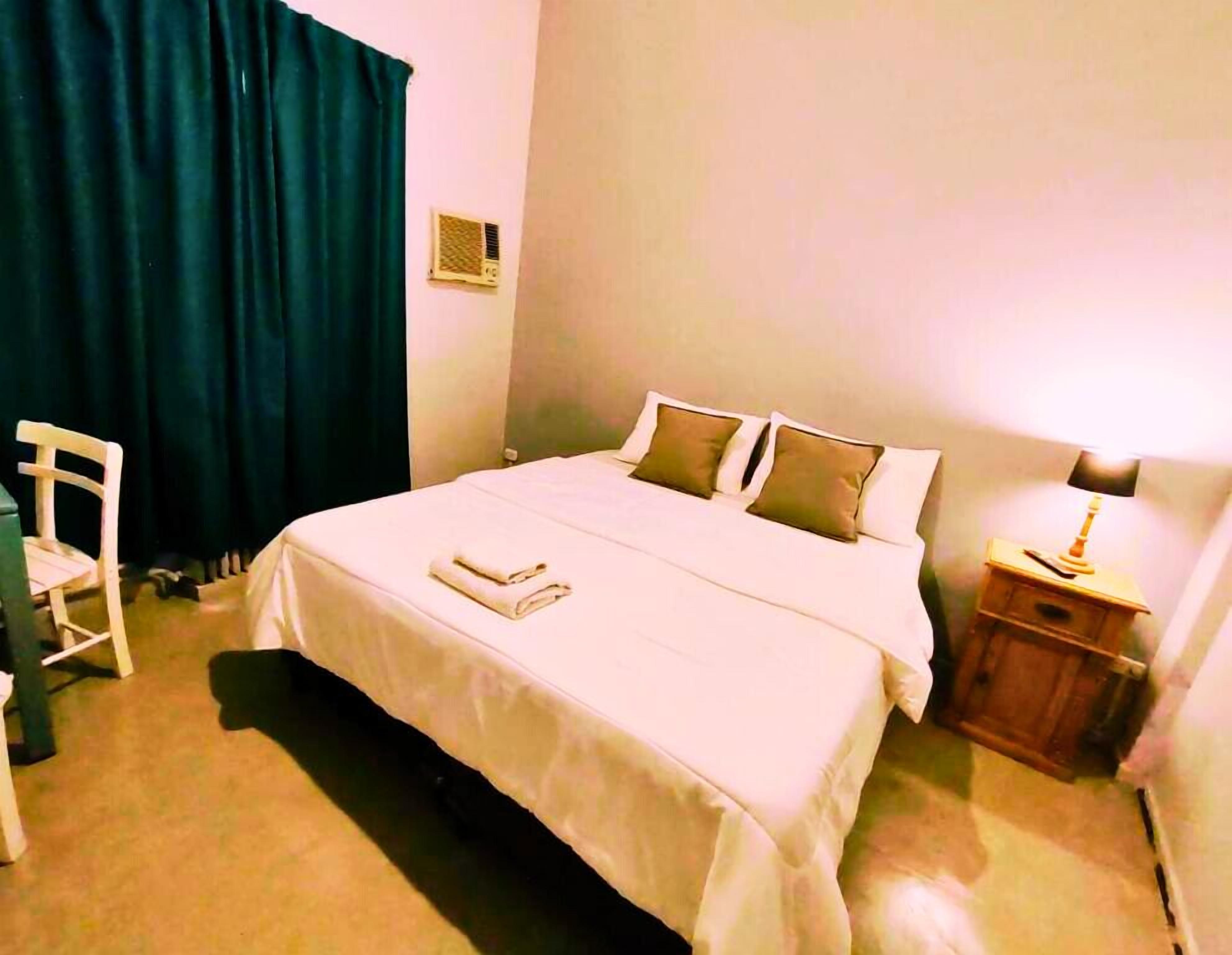 A hotel room with a bed, a bedside table with a lamp, a chair, and a window with curtains.