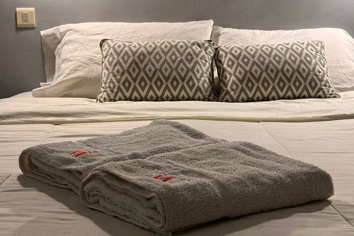 A neatly made bed with patterned pillows and a folded towel on a plain wall with a light fixture.