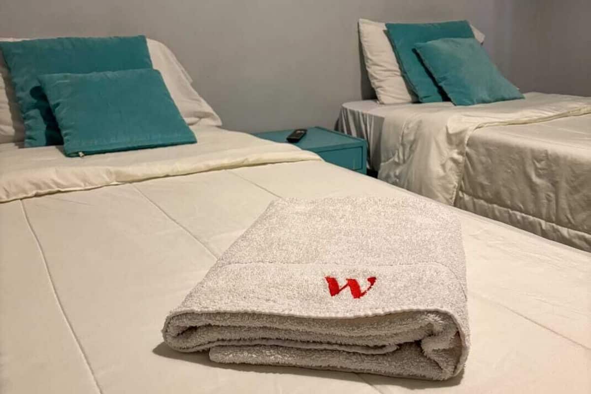 A hotel room with two beds, each with white bedding and teal pillows. A folded towel with a red 'W' logo is placed on the bed in the foreground.