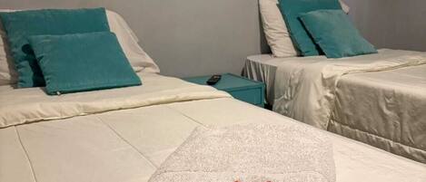 A hotel room with two beds, each with white bedding and teal pillows. A folded towel with a red 'W' logo is placed on the bed in the foreground.