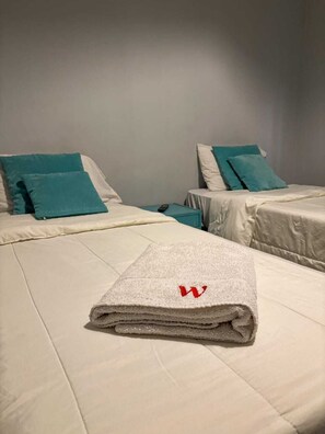 Une chambre d’hôtel avec deux lits, chacun agrémenté de draps blancs et d’oreillers turquoise. Une serviette pliée, ornée d’un logo « W » rouge, est posée sur le lit, au premier plan.