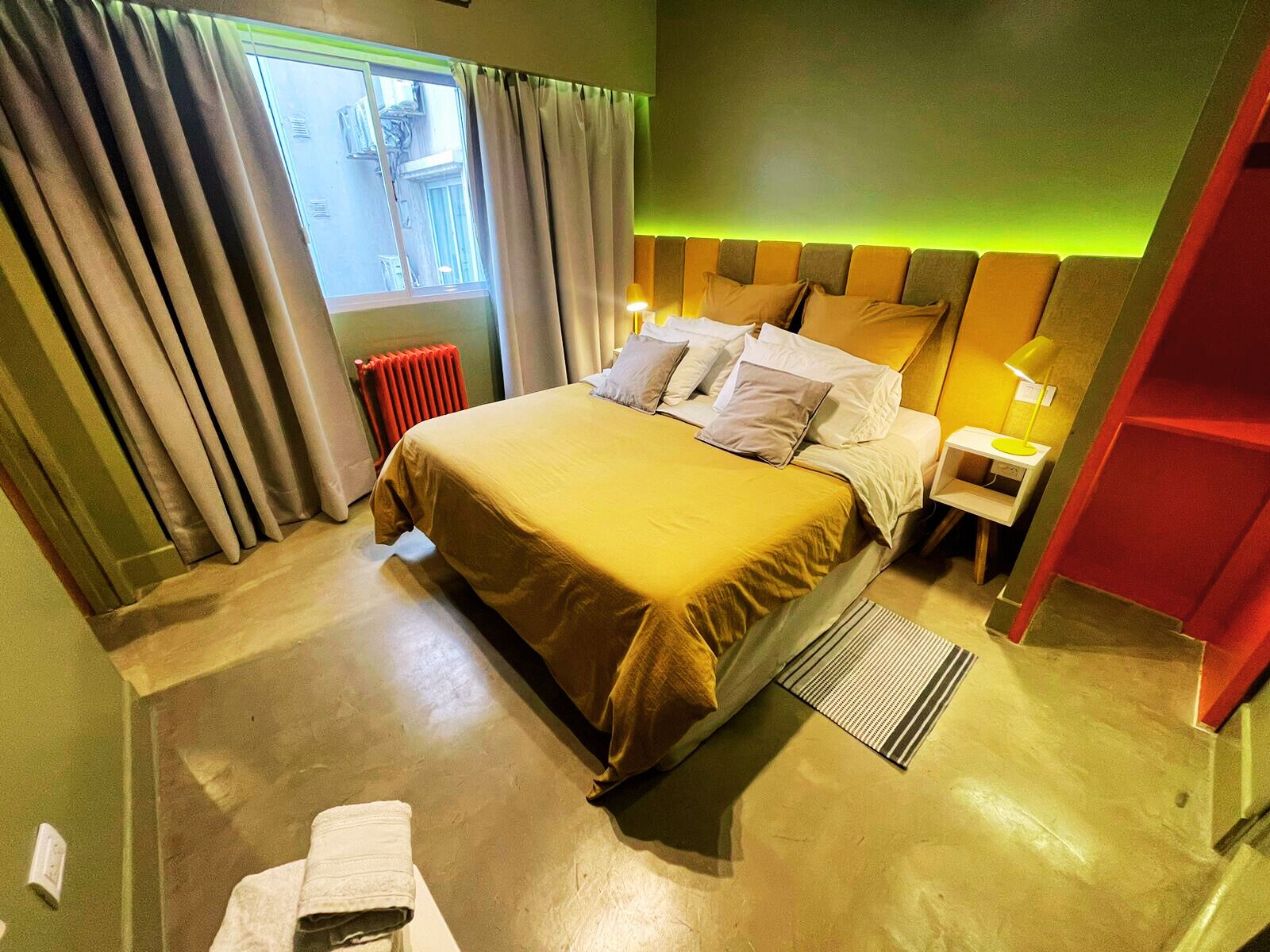 A hotel room with a large bed, a nightstand, a lamp, a radiator, and a window with curtains.