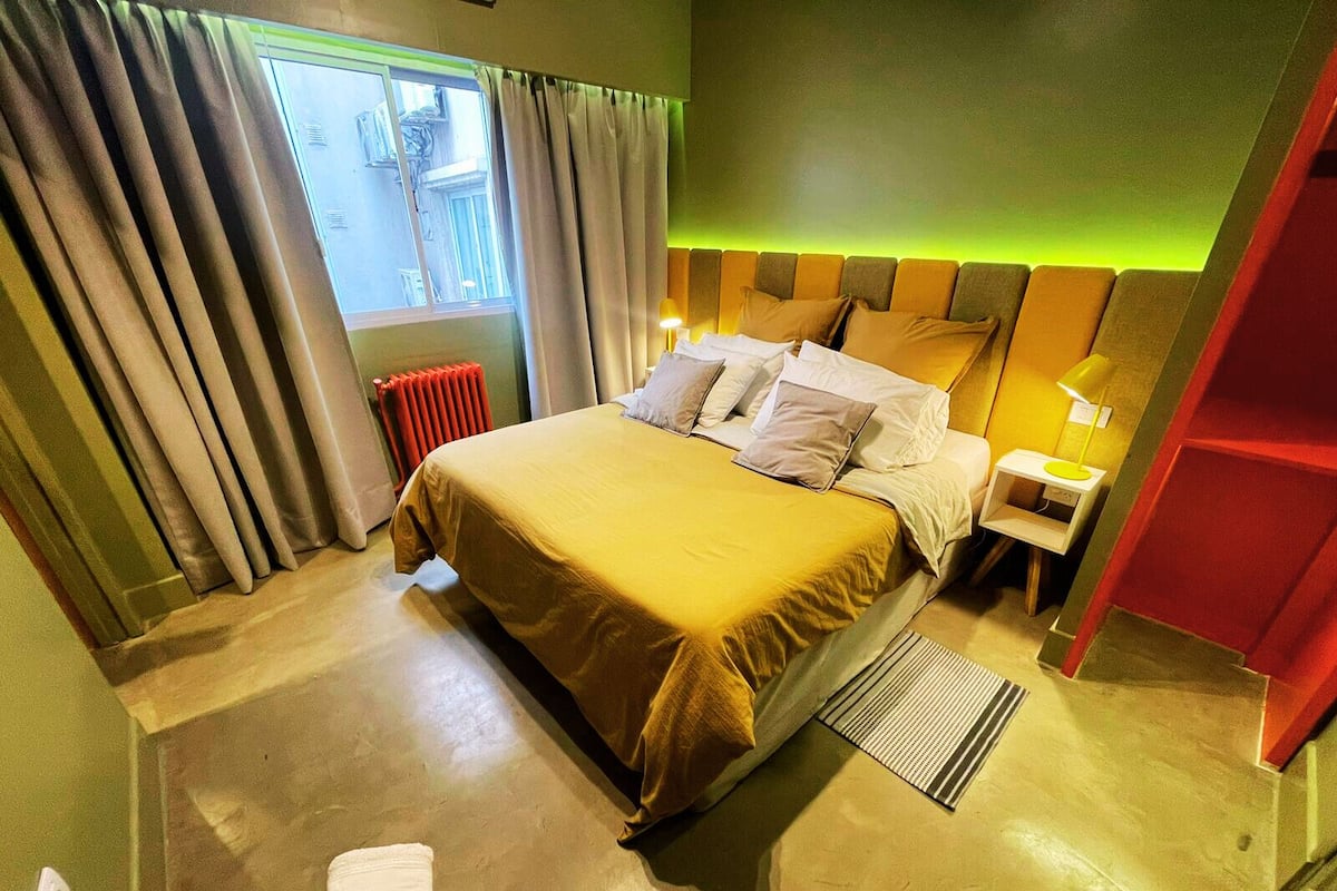 A hotel room with a large bed, a nightstand, a lamp, a radiator, and a window with curtains.